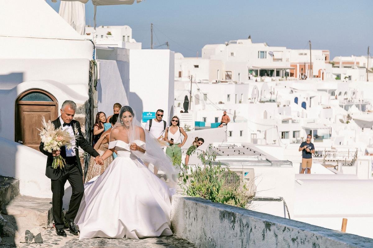 Bride walking through iconic whitewashed streets of Santorini, holding her dress with joy