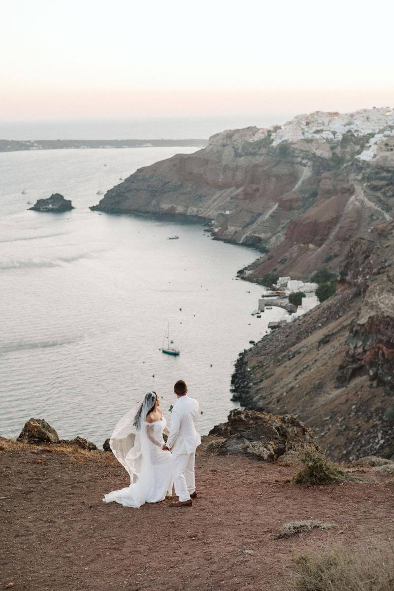 Bride and groom overlooking the sea from a dramatic cliffside in Santorini during sunset
